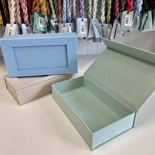 Open light green box with a closed blue box and beige box in the background, with yarn hanging above.