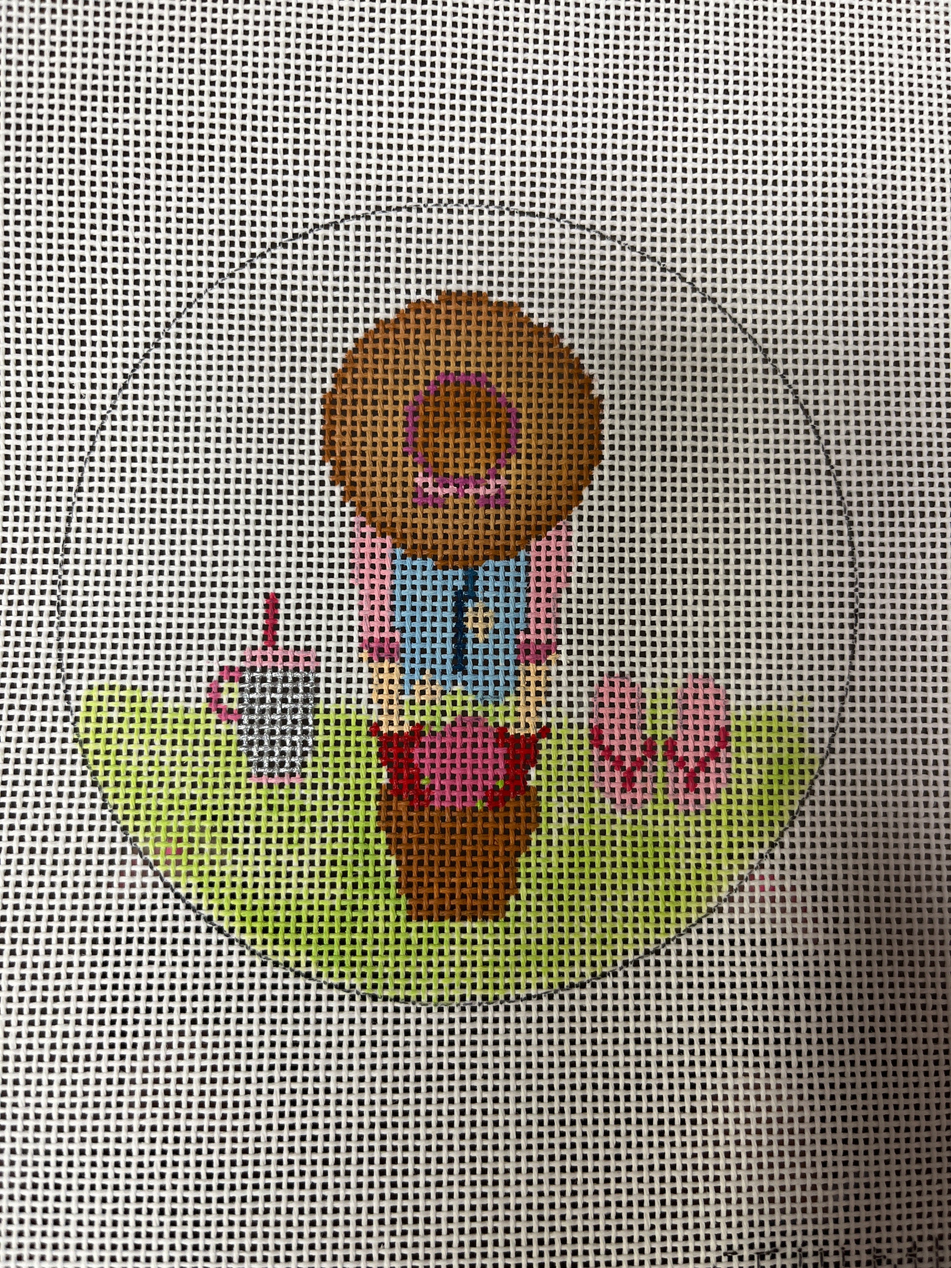 Cross-stitched design of a donut with a face, pink flowers, and a green base on a grid background.
