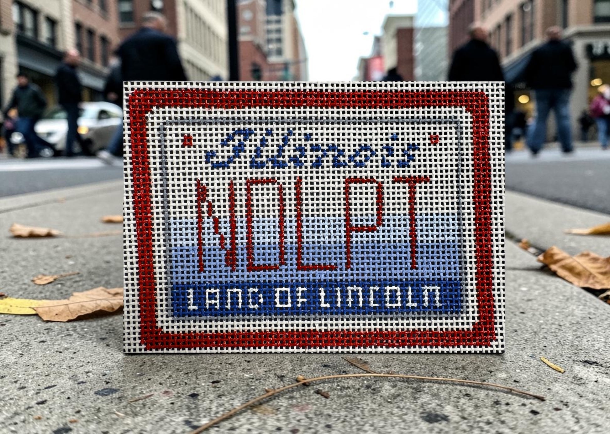 Small embroidered sign with 'NOLP' and 'Land of Lincoln' text on a city street background