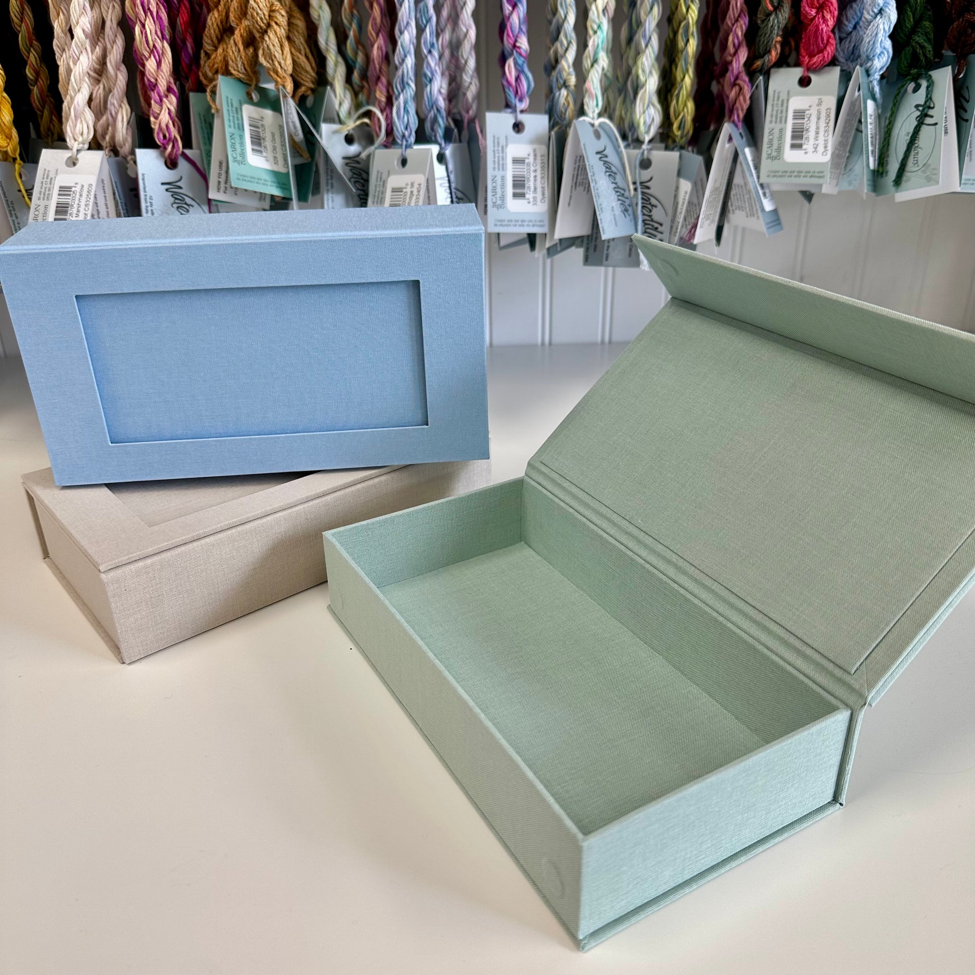 Open light green box with a closed blue box and beige box in the background, with yarn hanging above.