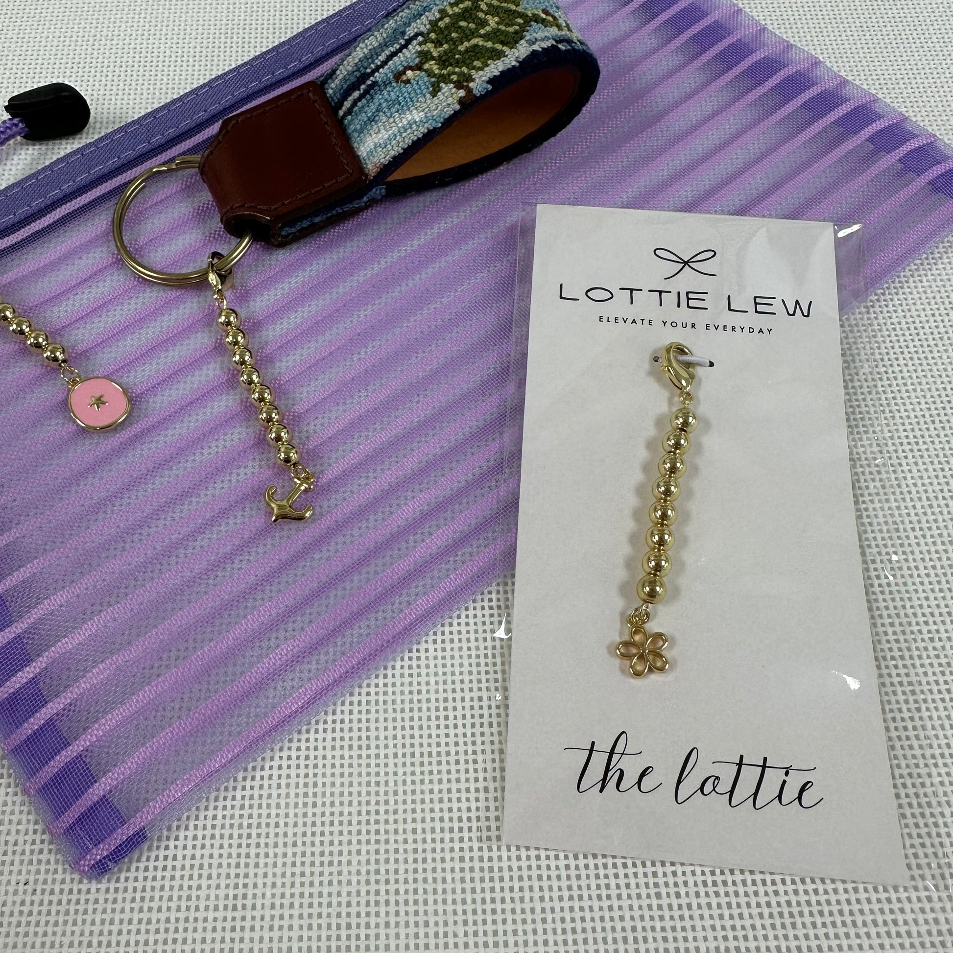 Gold beaded charm on a card with 'Lottie Lew' branding, next to a keychain and fabric strip.