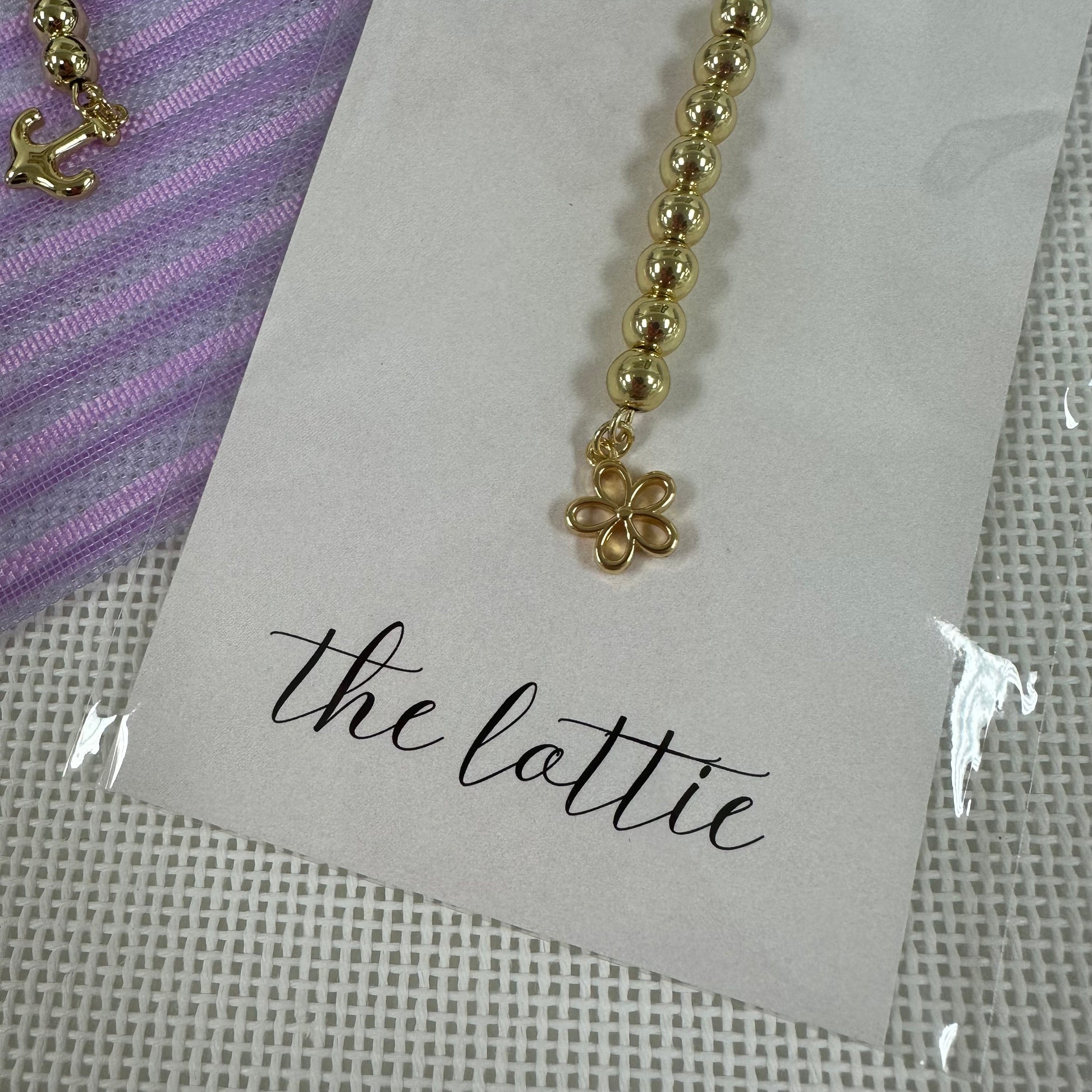 Gold beaded charm on a card with 'the lottie' branding, placed on a textured surface.