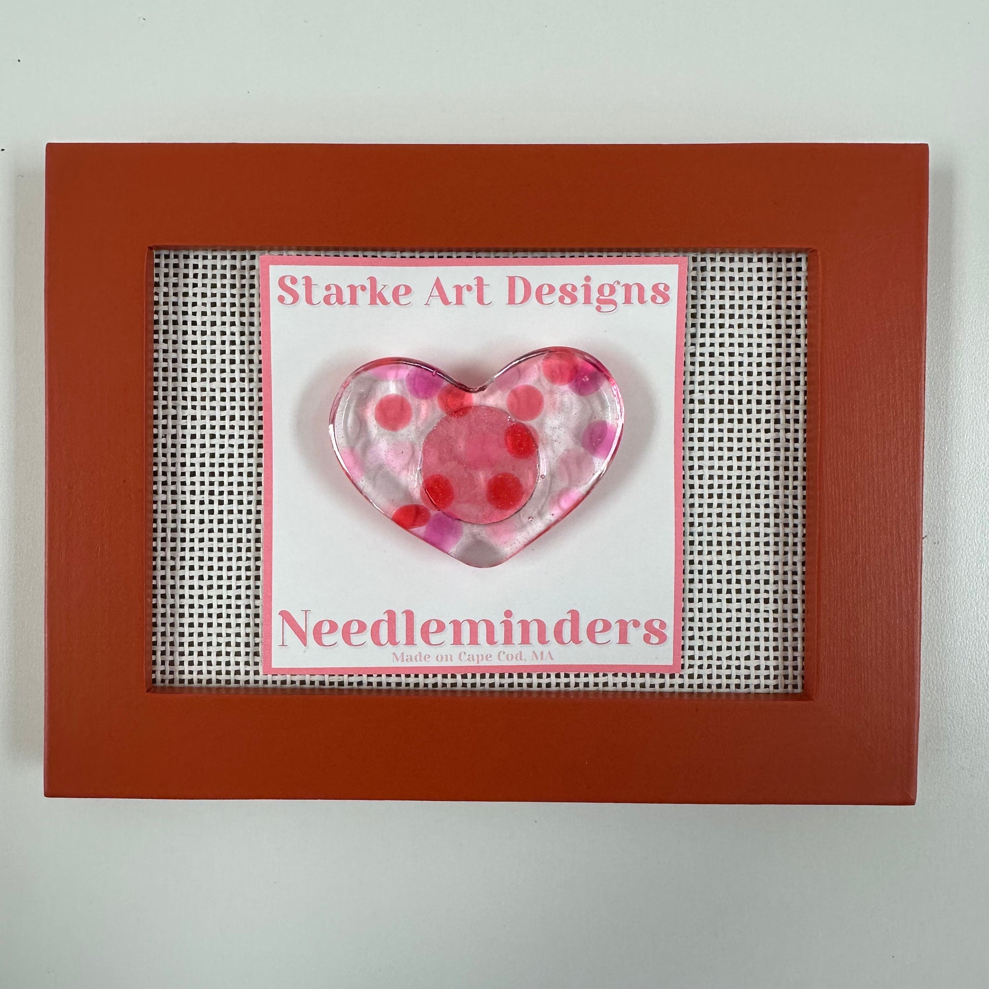 Heart-shaped pink and red polka dot needle minder with 'Starke Art Designs' branding in a brown frame.