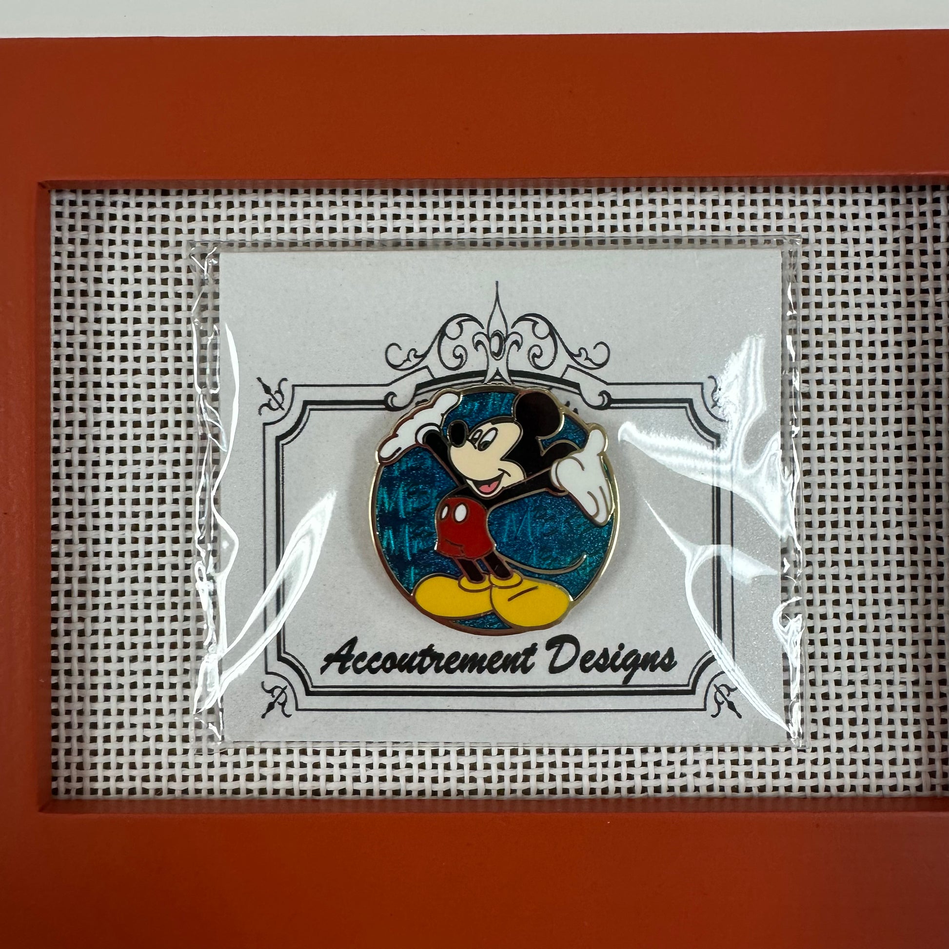 Pin featuring a character design by Accoutrement Designs in a clear plastic sleeve on a textured surface.