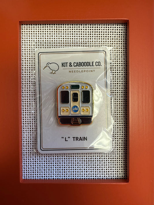 Pin of an 'L' train on a card with 'Kit & Caboodle Co.' branding, displayed against an orange background.