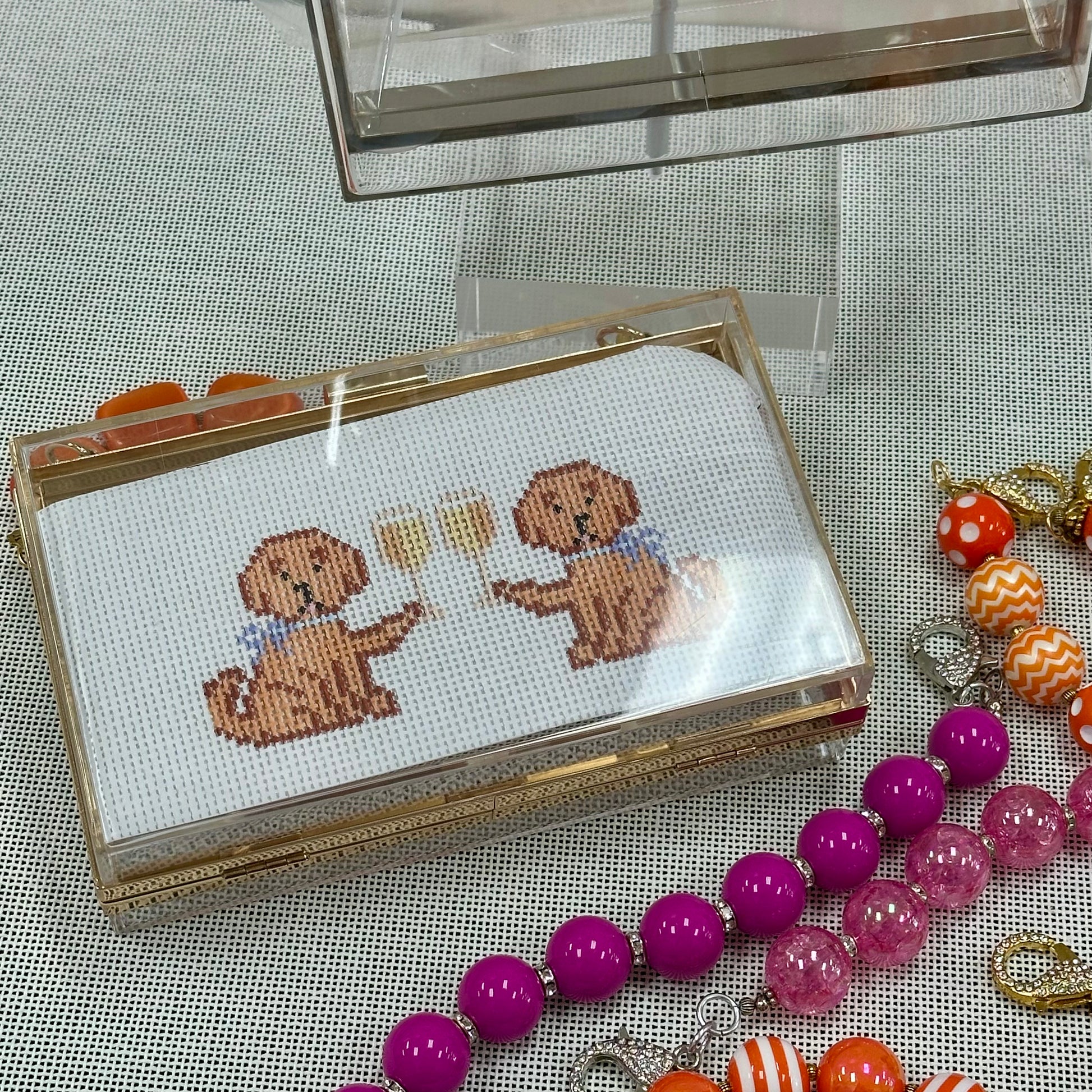Cross-stitched dogs in a clear box with colorful beads and jewelry on a textured surface