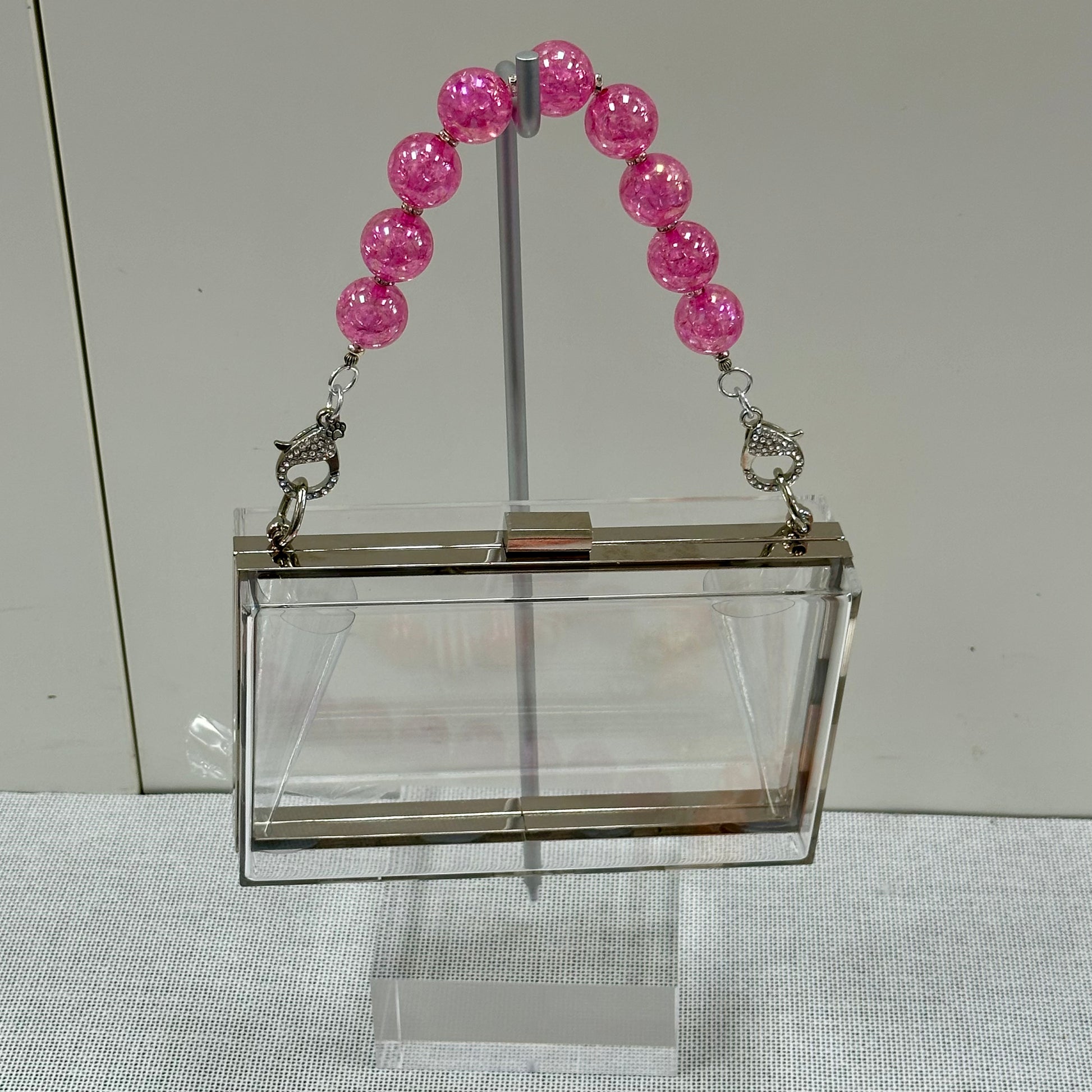 Clear rectangular handbag with pink beaded handle on a stand against a neutral background