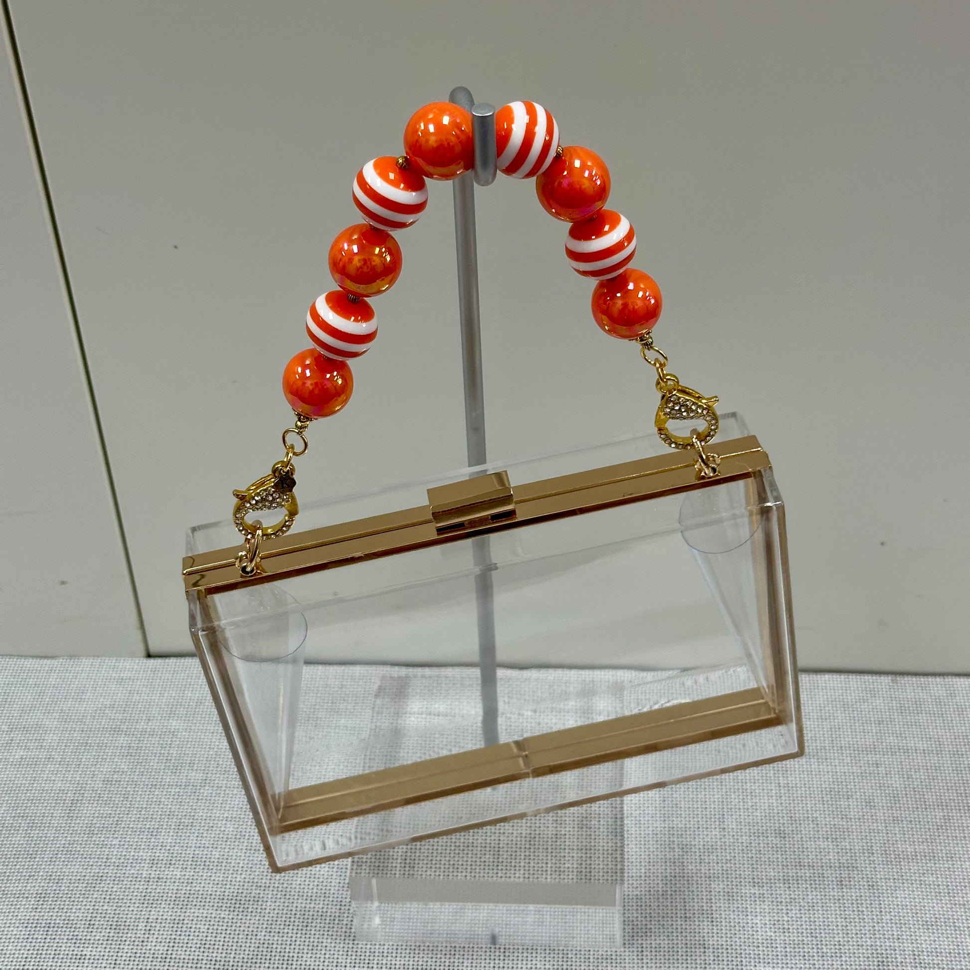 Clear handbag with gold accents and a bracelet with red and white striped beads on a gray surface.