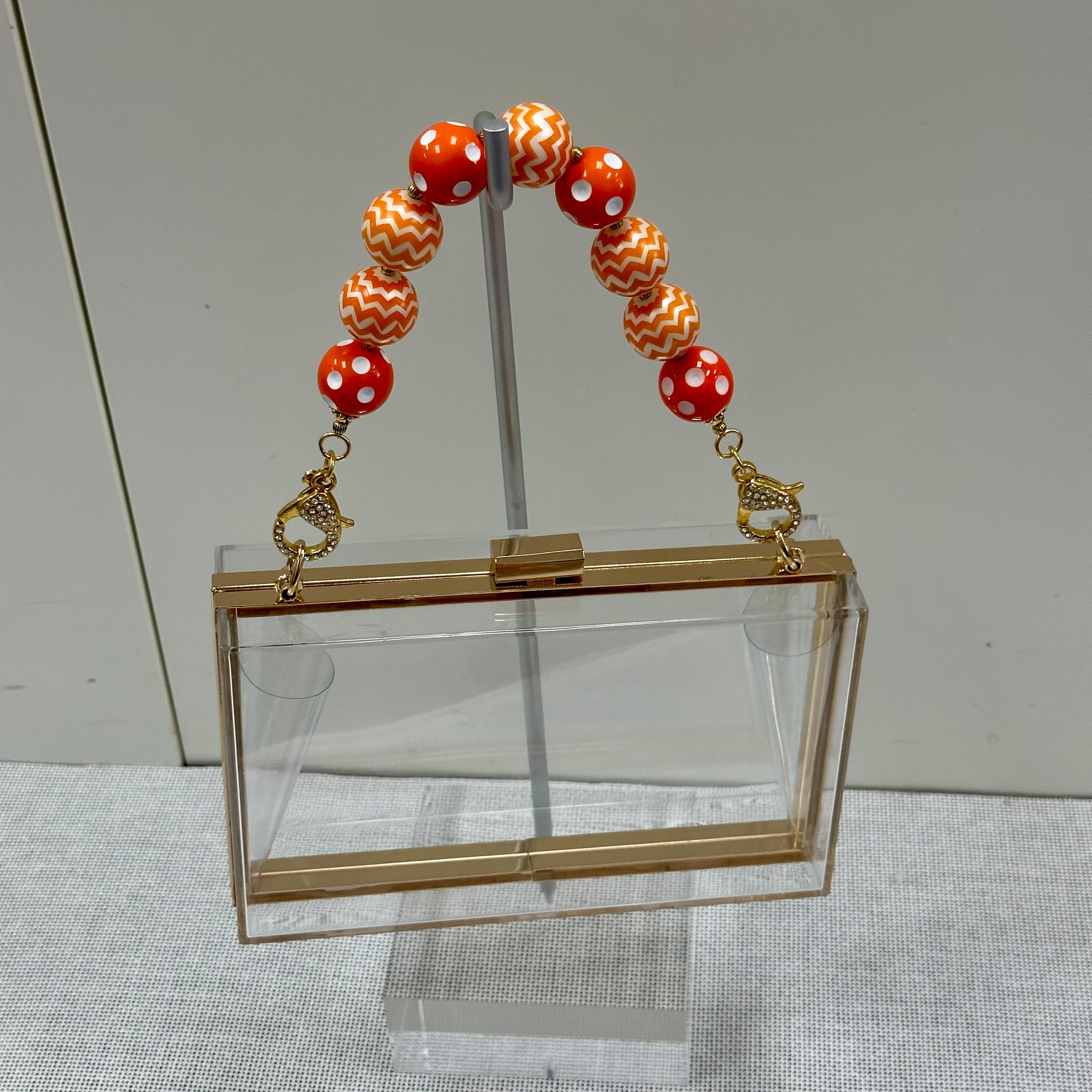 Clear handbag with gold accents and a bracelet on a stand against a neutral background