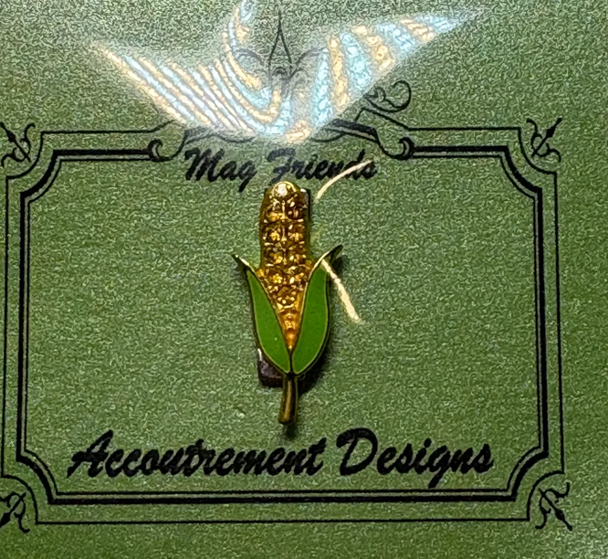 Corn on the cob pin with 'Accoutrement Designs' branding on a green background