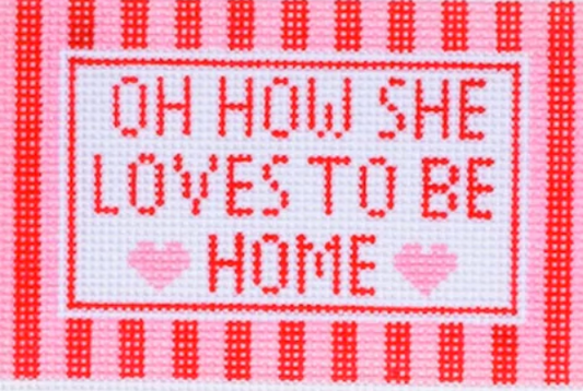 A square cross-stitch style design with the phrase “OH HOW SHE LOVES TO BE HOME”